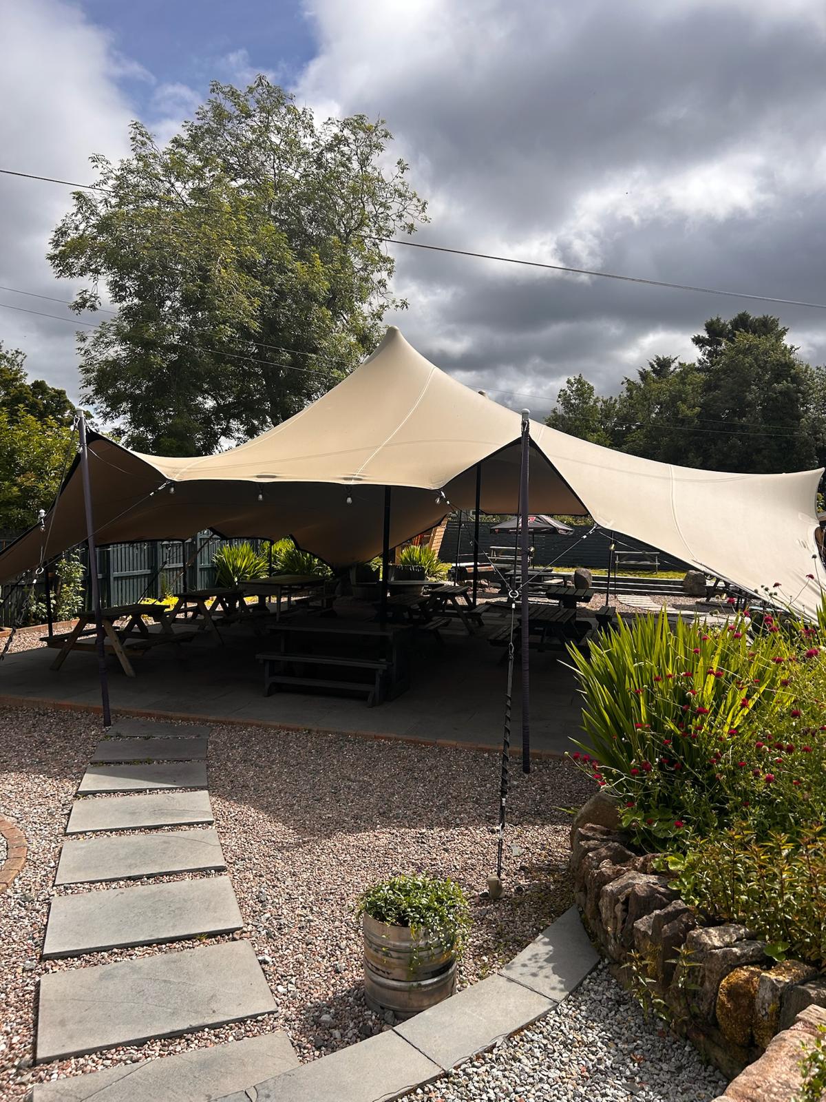 A stretch tent pitched over wooden picnic tables on the gravel garden patio